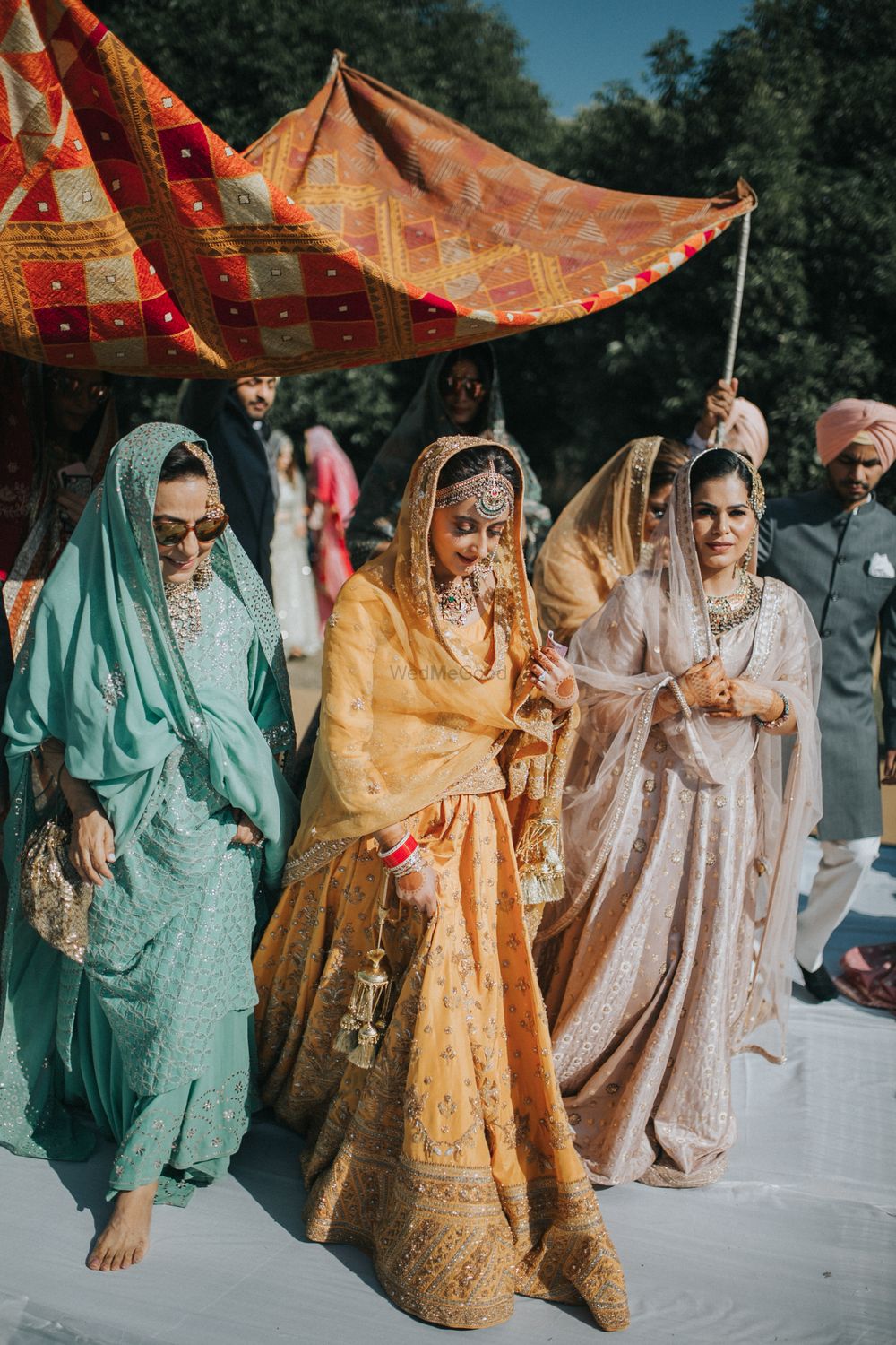 Photo of A bridal entry with bridesmaids and under a phulkari dupatta.