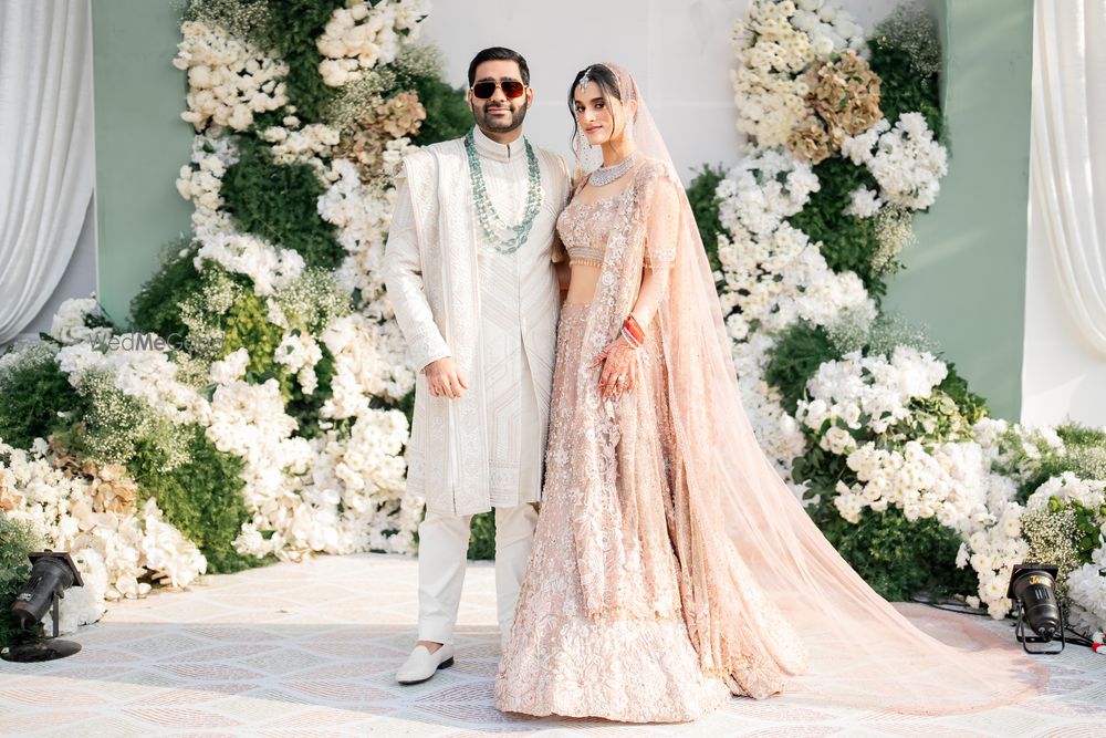 Photo of Couple portrait with gorgeous floral backdrop with the bride in a pastel peach lehenga and the groom in an all-white sherwani