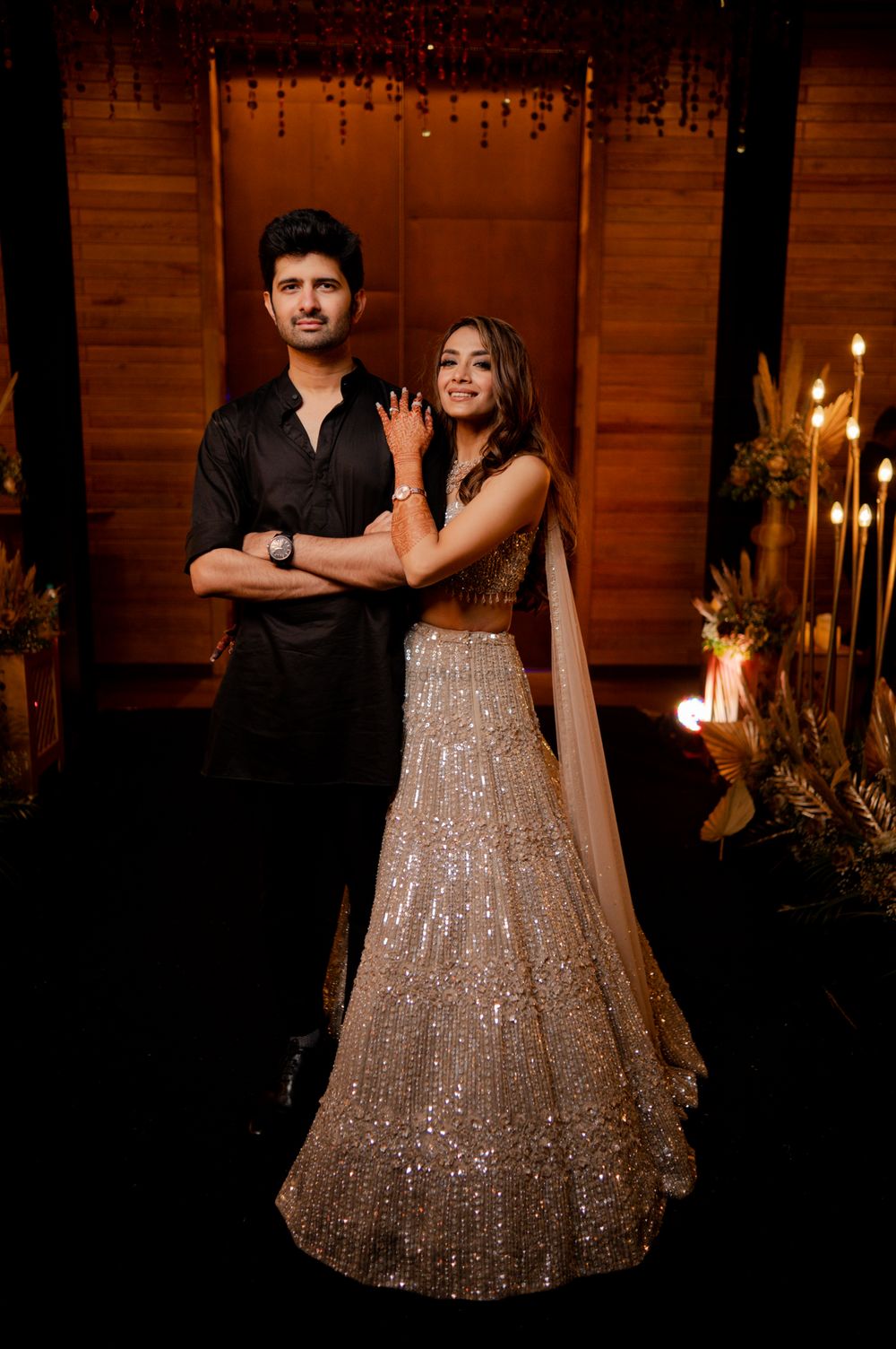 Photo of Couple portrait with the bride in a shimmery gold lehenga and groom in an all-black ensemble
