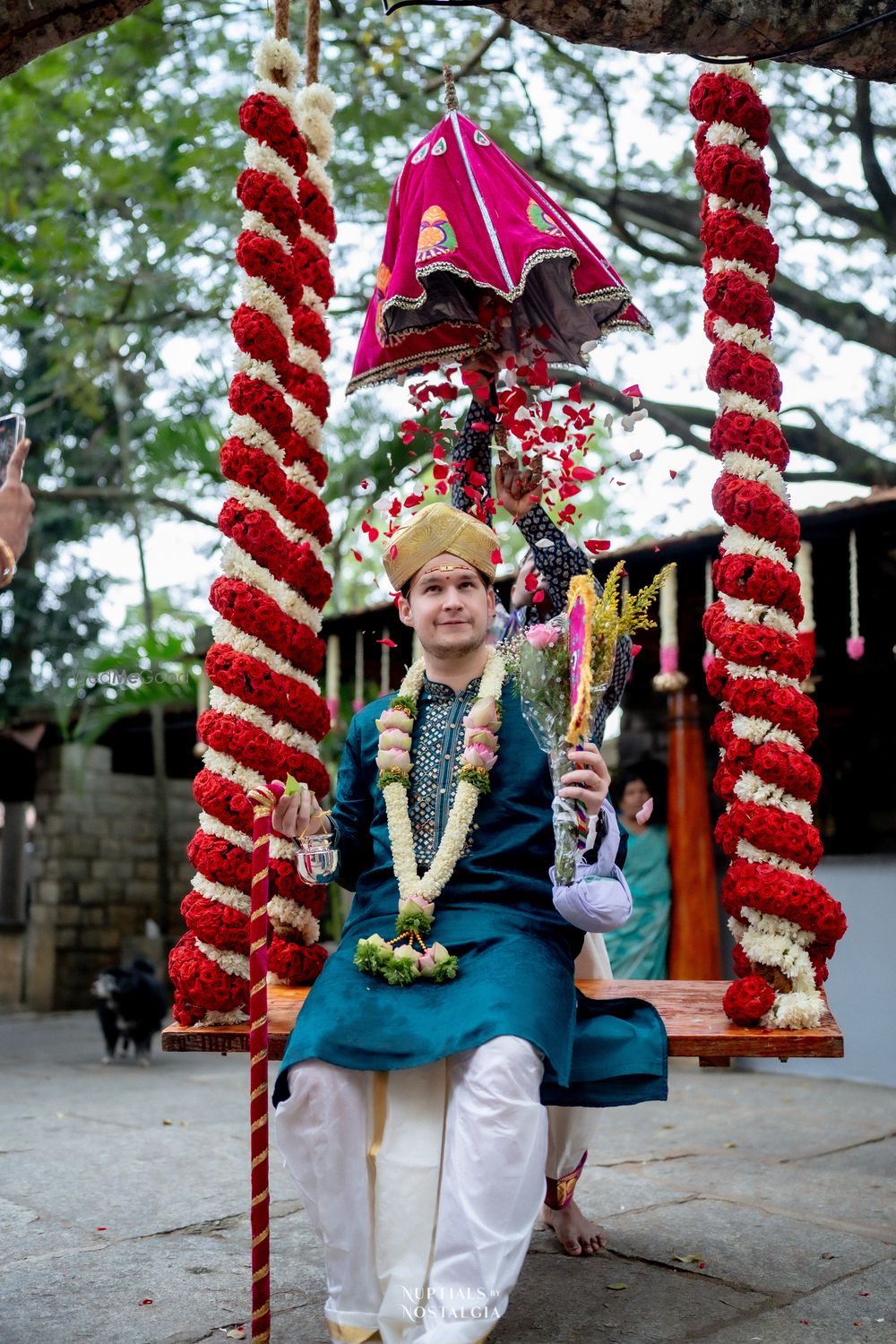 Photo of Groom to be sitting on a floral swing as an umbrella opens with flower shower