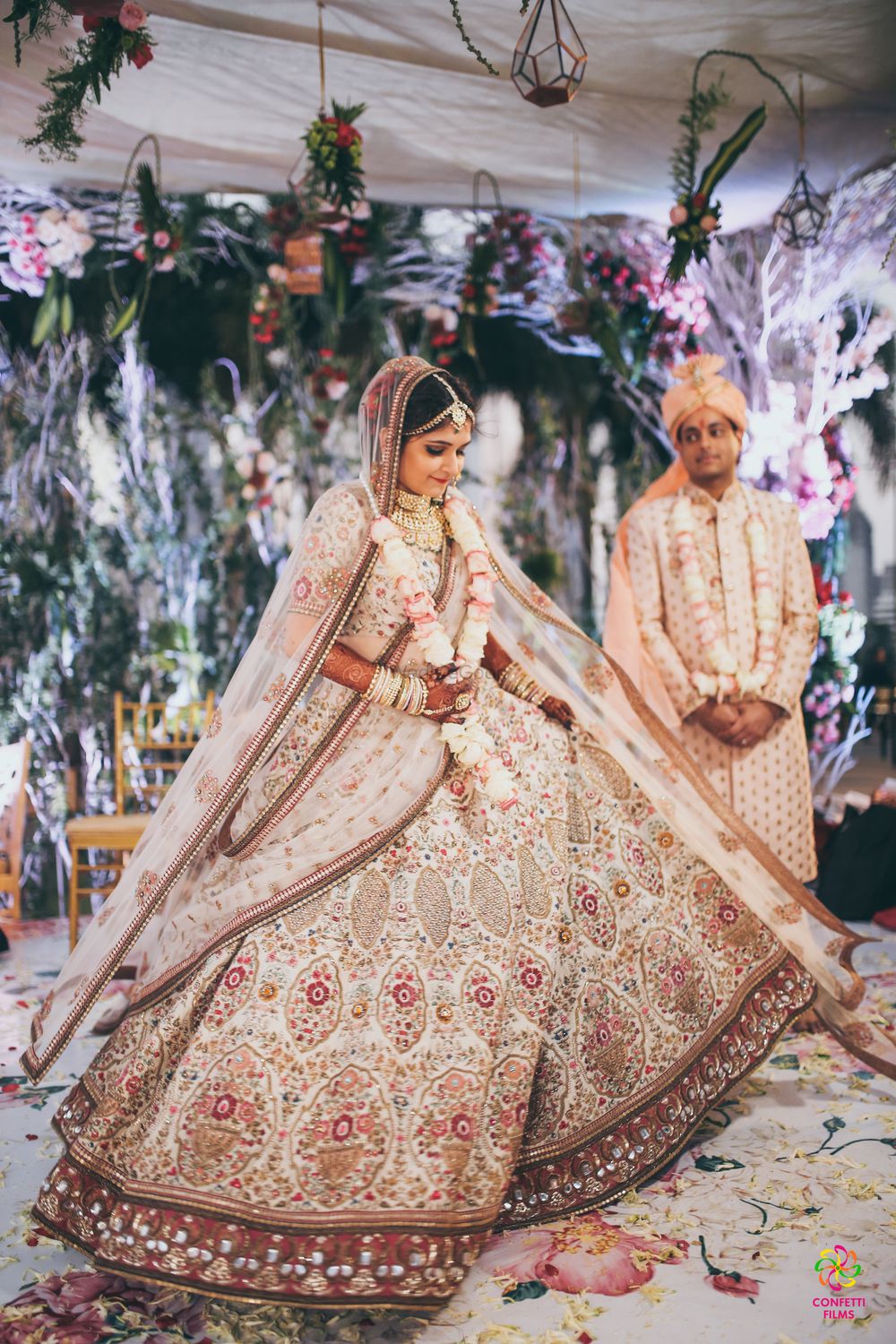Photo of A bride in creme lehenga twirls as her groom looks on