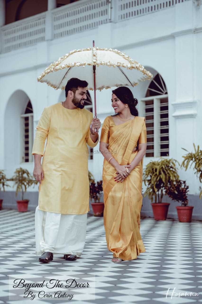Photo of A south Indian bride and groom in coordinated gold outfits with a floral umbrella