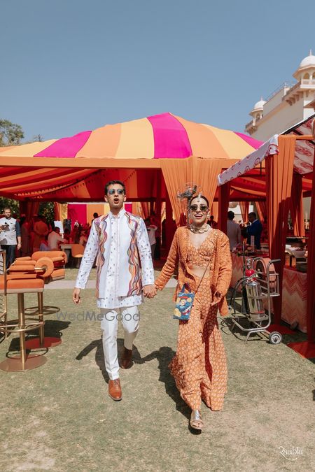 Photo of Fun shot of the couple on the mehendi day with the bride in an orange outfit and funky Lays bag