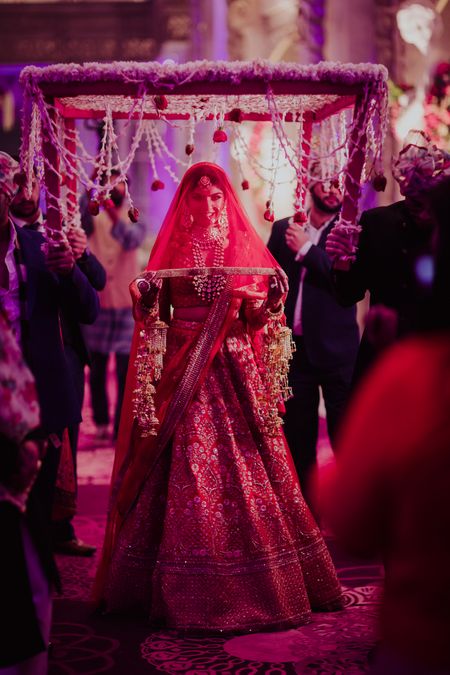 A bride in a red lehenga and a veil entering under a phoolon ki chaadar