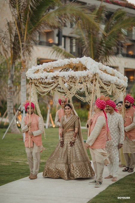 A bride entering under a gorgeous phoolon ki chaadar with tassels and white flowers 