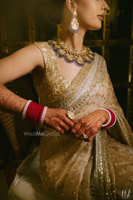 Photo of Gorgeous shot of the bride wearing a statement bib style necklace with lavender detailing, statement rings and a red chura to compliment her gold saree look.