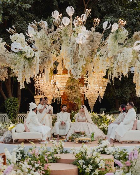 Photo of Super royal all-white mandap decor with florals and chandeliers, with female priestess conducting the wedding