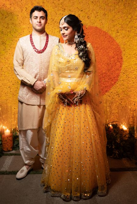 A bride in a yellow lehenga posing infront of a wall decorated with marigold flowers with her groom to be!