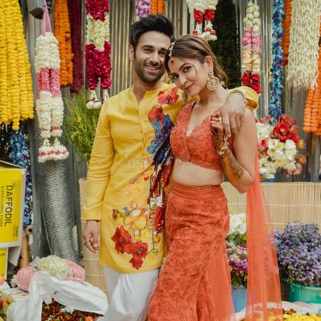 Couple portrait with groom in a yellow floral kurta and bride in a bright orange co-ord set with cape style dupatta for the haldi