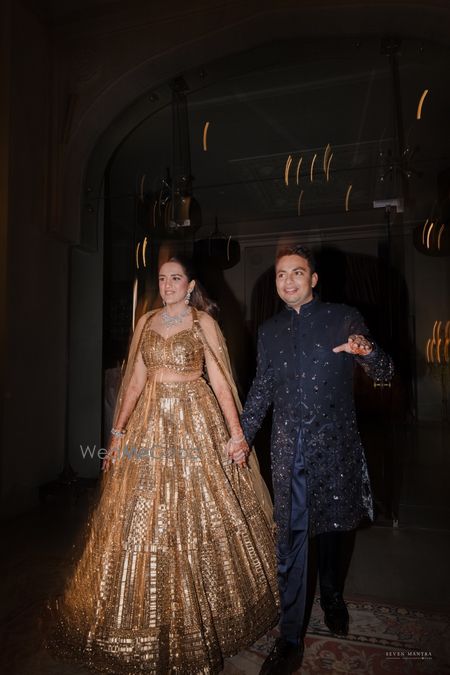 Couple entering their sangeet event with the bride in a gold lehenga with cape dupatta and groom in a navy blue sherwani