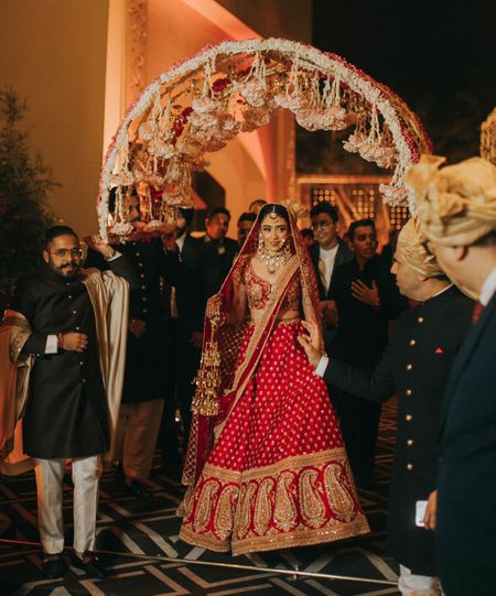 Photo of bride in red lehenga entering under a circular phoolon ka chadar