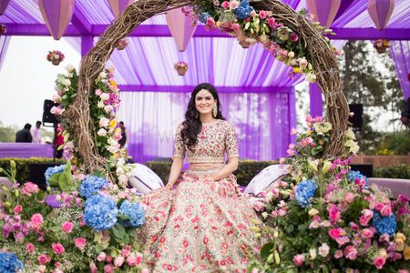 Photo of A bride sitting under a wreath of flowers in a nude peach lehenga for her mehendi.
