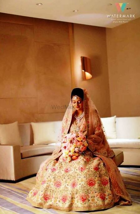 A bride in a floral lehenga sitting with a flower bouquet