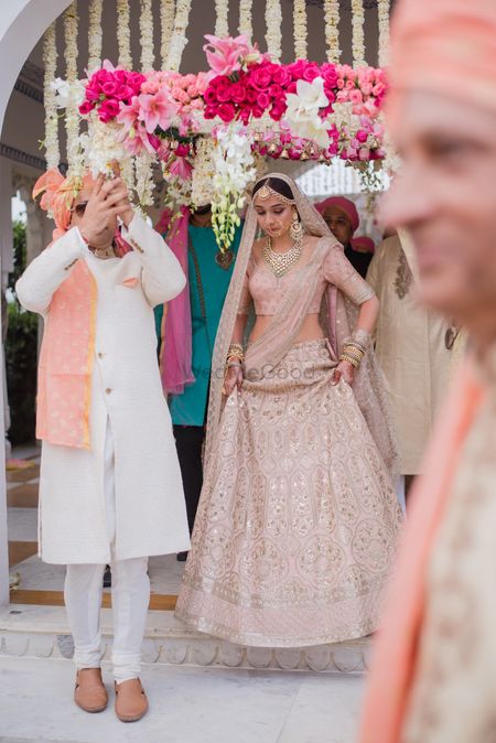 A bride in a soft pink lehenga entering under a phoolon ki chaadar