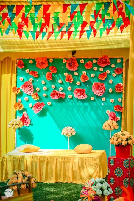 Photo of Yellow Mehendi seating with a backdrop decorated with paper flowers.